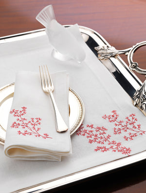 White linen napkin and placemat with coral red embroidery, styled with silver flatware and a decorative glass bird on a polished serving tray – luxury coastal table linens by Plein Soleil.