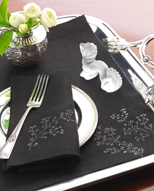 Black linen placemat and napkin set embroidered with delicate white branches, styled with silver flatware, floral arrangement, and crystal sea horses – elegant Mediterranean-inspired table linens by Plein Soleil.