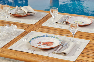 Elegant poolside table setting with knot-embroidered linen placemats, artisanal ceramic plates, crystal glassware, and fine silverware – luxury outdoor dining linens by Plein Soleil.