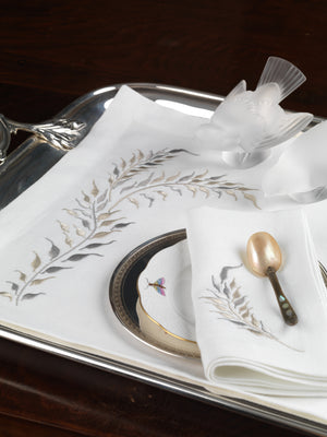 White linen placemat and napkin set with soft taupe leaf embroidery, styled with antique silver tray, porcelain plate, and gold spoon – heirloom-quality embroidered table linens by Plein Soleil.