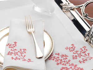 White linen napkin and placemat with coral red embroidery, styled with silver flatware and a decorative glass bird on a polished serving tray – luxury coastal table linens by Plein Soleil.