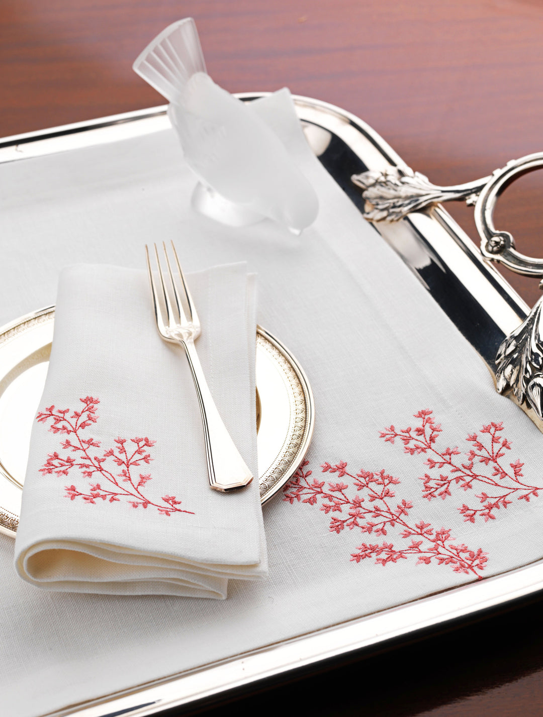 White linen napkin and placemat with coral red embroidery, styled with silver flatware and a decorative glass bird on a polished serving tray – luxury coastal table linens by Plein Soleil.
