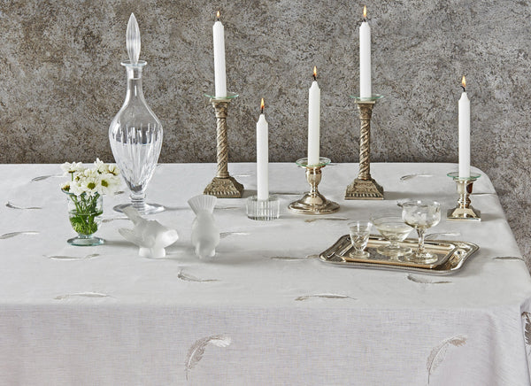 Elegant white linen tablecloth with feather embroidery, styled with crystal glassware, brass candlesticks, and fresh flowers – artisanal European table linens by Plein Soleil.
