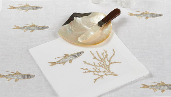 White linen placemat with gold and silver embroidered fish and coral, styled with mother-of-pearl knife and crystal glassware – luxury coastal table linens by Plein Soleil.
