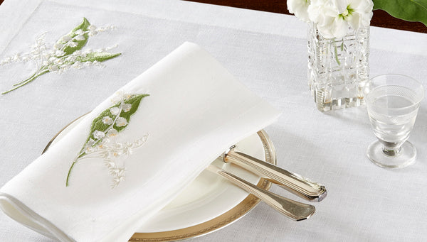 White linen napkin with lily embroidery, styled with fine porcelain, silver cutlery, and crystal glassware – luxury table linens by Plein Soleil.