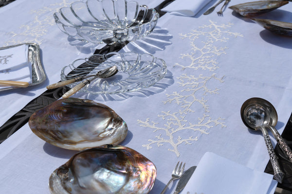 Dining table setting with shell plates, cutlery, and glassware on a white tablecloth with gold leaf design.