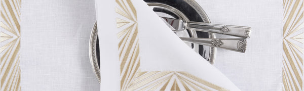 Elegant white linen napkin with sunburst gold embroidery on a silver-rimmed plate with vintage cutlery — luxury table setting by Plein Soleil.