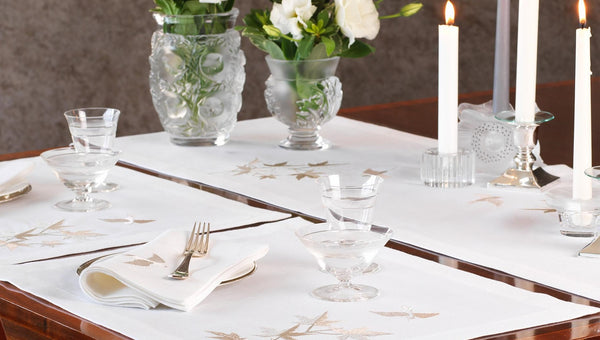 Elegant white linen table runner and napkins embroidered with swallows and maple leaves, styled with crystal glassware, candles, and floral arrangements – bespoke nature-inspired table linens by Plein Soleil.
