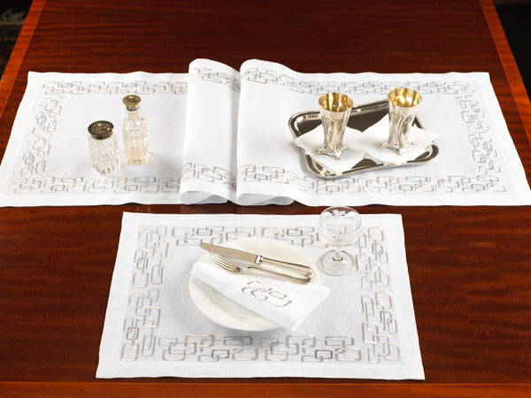 White linen placemat and table runner with taupe chain embroidery, styled with silverware, crystal glassware, and gold-lined goblets – handcrafted luxury table linens by Plein Soleil.