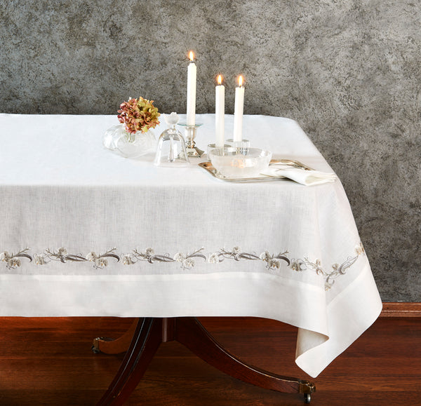Refined dining setup featuring a white linen tablecloth with floral ribbon embroidery, candles, and glassware — bespoke B2B linens by Plein Soleil.